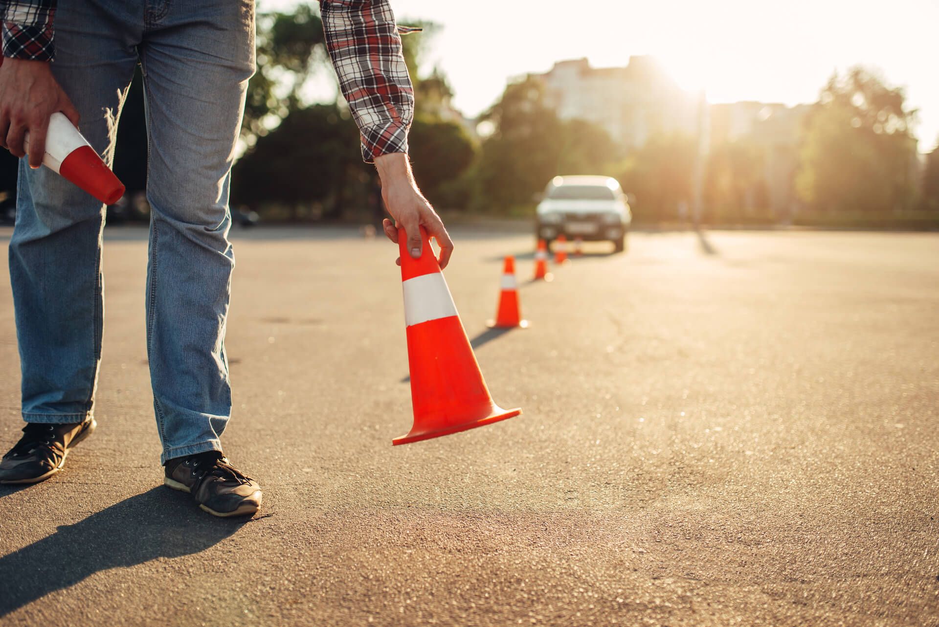 Eine Person stellt orangefarbene Verkehrskegel auf einer sonnenbeschienenen Straße mit einem Auto im Hintergrund auf.